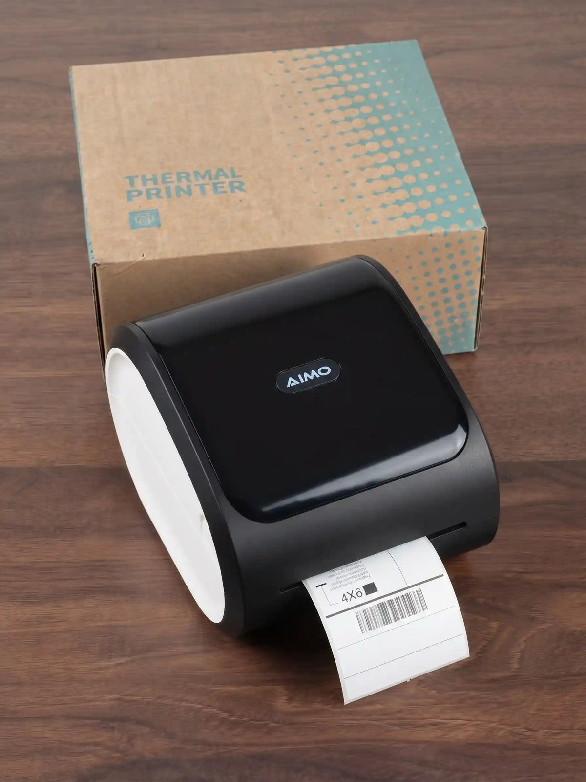 A black AIMO thermal label printer with a white roll of labels partially unspooled, placed on a dark wood surface in front of its brown cardboard packaging box.