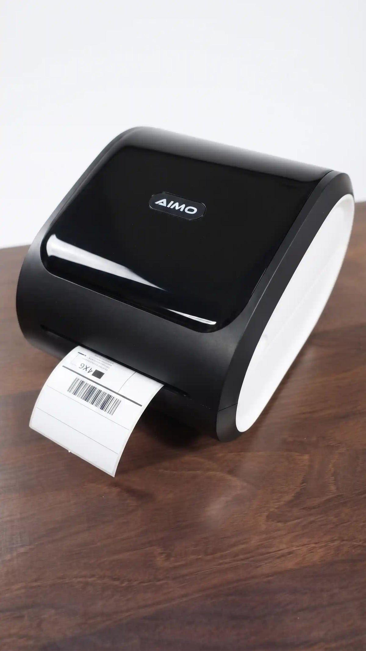 A black-and-white AIMO thermal label printer on a dark wooden table, with a white label partially ejected.